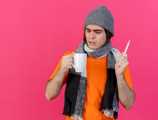Hombre enfermo joven confundido con sombrero de invierno con bufanda sosteniendo el termómetro mirando una taza de té en su mano aislado sobre fondo rosa