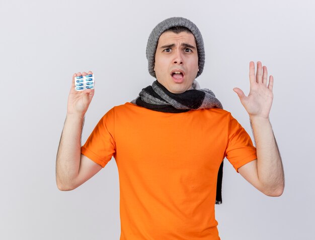 Hombre enfermo joven insatisfecho con sombrero de invierno con bufanda sosteniendo pastillas y mostrando gesto de parada aislado en blanco