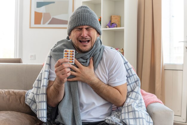 Hombre enfermo llorando con bufanda alrededor del cuello con gorro de invierno sosteniendo un blister de medicina y poniendo la mano en el pecho sentado en el sofá en la sala de estar