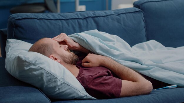 Hombre enfermo mirando el termómetro para comprobar la medición de la fiebre mientras duerme. Persona enferma que sostiene un instrumento médico que mide la temperatura y descansa en una manta. Adulto enfermo con enfermedad