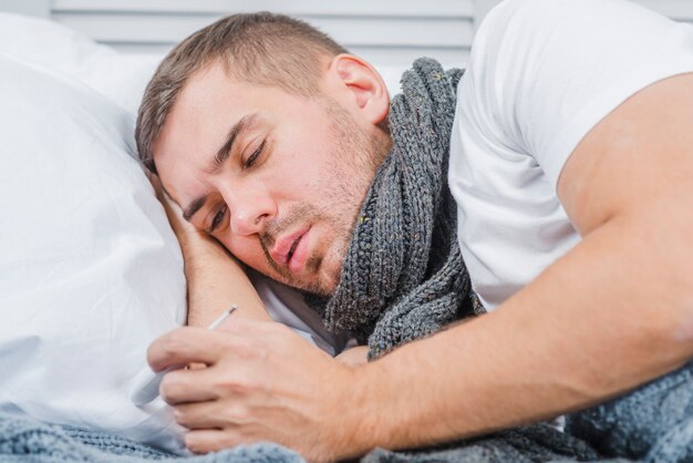 Hombre enfermo que lleva una bufanda gruesa alrededor del cuello mirando el termómetro