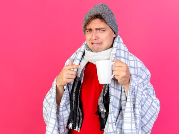 Hombre enfermo rubio guapo joven irritado con sombrero de invierno y bufanda envuelto en cuadros sosteniendo y apuntando a la taza aislada en la pared carmesí con espacio de copia