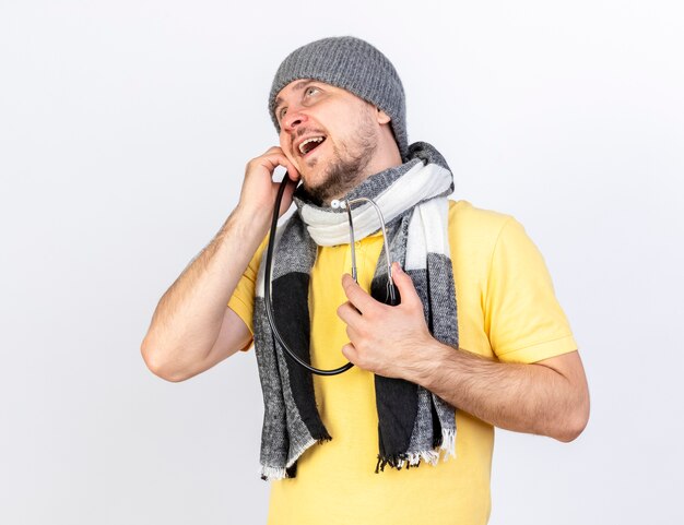 Hombre enfermo rubio joven complacido con bufanda y sombrero de invierno sostiene estetoscopio mirando hacia arriba aislado en la pared blanca