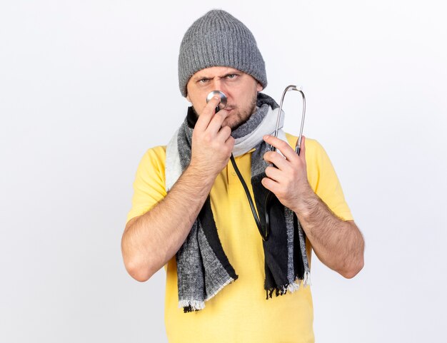 Foto gratuita hombre enfermo rubio joven disgustado con bufanda y sombrero de invierno sostiene estetoscopio aislado en la pared blanca