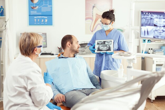 Hombre enfermo sentado en el sillón estomatológico escuchando al médico mientras mira la tableta en la clínica dental