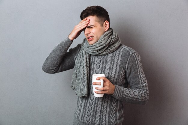 Hombre enfermo en suéter y bufanda con dolor de cabeza mientras sostiene una taza de té