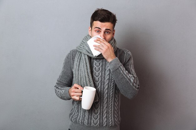 Hombre enfermo en suéter y bufanda con secreción nasal mientras sostiene una taza de té y mira