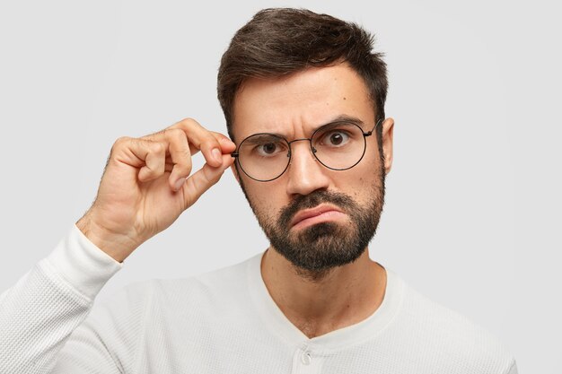 Hombre enojado sin afeitar mira seriamente a la cámara, frunce el ceño, toca el borde de las gafas, ve desconcertado