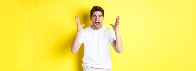 Hombre enojado gritando y dándose la mano haciendo una mueca de odio de pie odioso contra el fondo amarillo