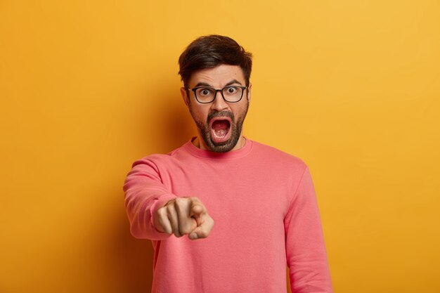 Hombre enojado indignado indica, te culpa, grita de irritación, dice que eres culpable, usa anteojos y suéter rosado, posa contra la pared amarilla, regaña a la persona por cometer un error