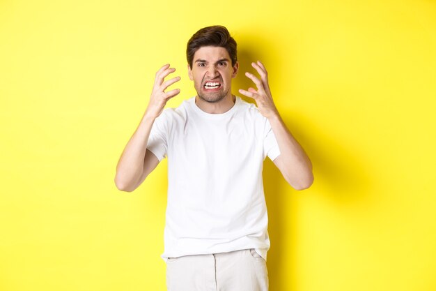 Hombre enojado que parece enojado, haciendo muecas y dándose la mano furioso, de pie indignado contra el fondo amarillo.