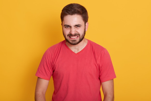 El hombre enojado que vestía una camiseta casual roja, disgustado, mantiene las manos tensas, el tipo sin afeitar con elegante peinado torció la cara con ira, posando aislado en amarillo.