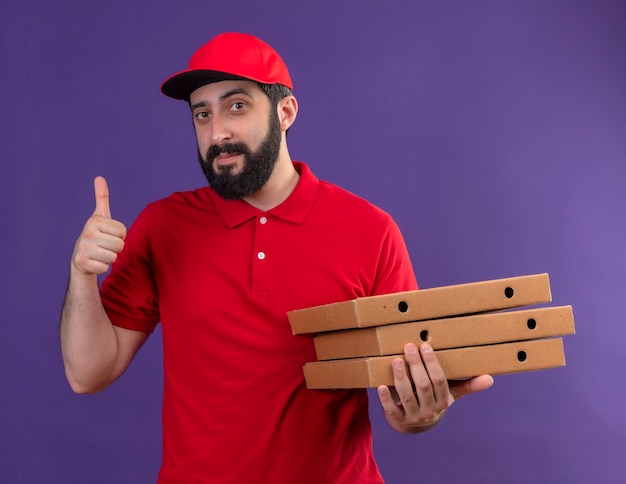 Foto gratuita hombre de entrega apuesto joven complacido con uniforme rojo y gorra sosteniendo cajas de pizza y mostrando el pulgar hacia arriba aislado en la pared púrpura