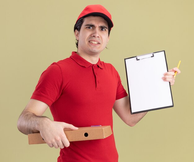 Hombre de entrega caucásico joven con el ceño fruncido en uniforme rojo y gorra sosteniendo el paquete de pizza que muestra el portapapeles con un lápiz en la mano