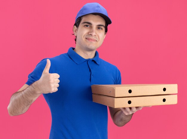 Hombre de entrega caucásico joven complacido en uniforme azul y gorra sosteniendo paquetes de pizza mostrando el pulgar hacia arriba