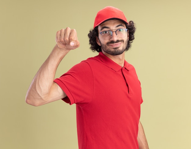 Hombre de entrega caucásico joven complacido en uniforme rojo y gorra con gafas haciendo gesto de golpe