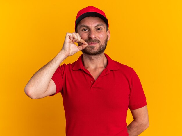 Hombre de entrega caucásico joven complacido en uniforme rojo y gorra manteniendo la mano detrás de la espalda cerrando la boca