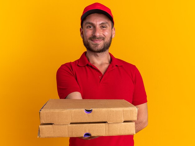 Hombre de entrega caucásico joven complacido en uniforme rojo y gorra manteniendo la mano detrás de la espalda mirando a la cámara estirando paquetes de pizza hacia la cámara aislada en la pared naranja