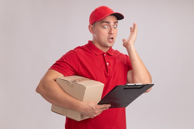 Hombre de entrega joven asustado vestido con uniforme con gorra sosteniendo la caja y mirando el portapapeles en la mano aislada en la pared blanca