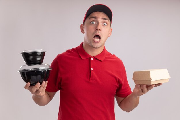 Hombre de entrega joven asustado vestido con uniforme con gorra sosteniendo el paquete de comida de papel con recipiente de comida aislado en la pared blanca