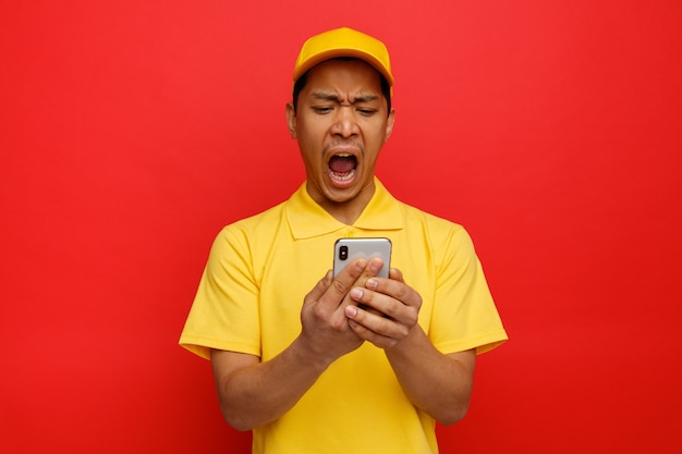 Hombre de entrega joven estresado con gorra y uniforme sosteniendo y mirando el teléfono móvil gritando