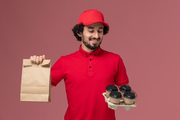 Hombre de entrega de mensajería masculino de vista frontal en camisa roja y capa sosteniendo tazas de café de entrega marrón con paquete de comida en el trabajo de empleado de entrega de servicio de pared rosa