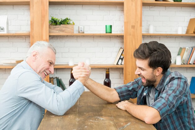 Hombre envejecido y chico joven con las manos juntas en el desafío de la lucha de brazos en la mesa en la habitación