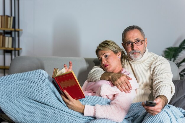 Hombre envejecido con control remoto de TV viendo televisión y libro de lectura de mujer en el sofá