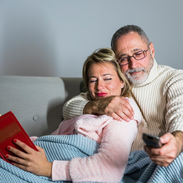 Hombre envejecido con control remoto de TV viendo televisión y mujer alegre con libro en sofá