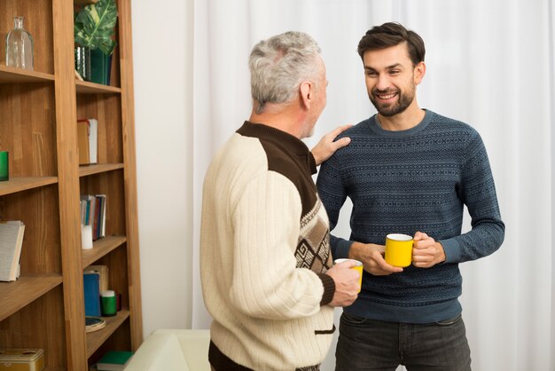 Hombre envejecido con la mano en el hombro del joven sonriente con tazas en la habitación