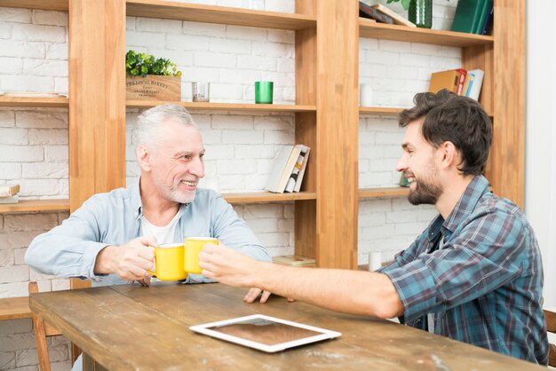 Hombre envejecido sonriente que golpea las tazas con el chico joven feliz en la mesa con tableta