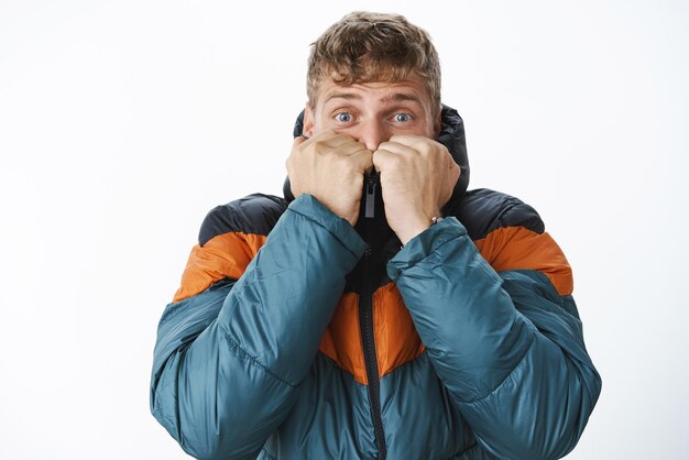 Hombre escondiéndose del invierno y la nieve con una chaqueta acolchada tirando del cuello en la cara levantando las cejas y con los ojos muy abiertos asustados por el calentamiento del clima frío mientras compran un abrigo fresco al aire libre para la temperatura de congelación