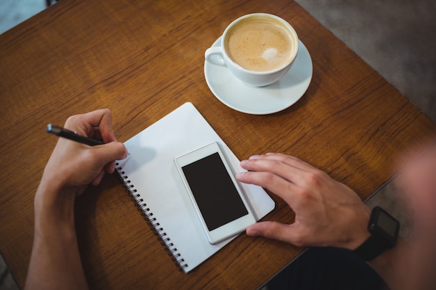 El hombre escribe en la libreta durante el uso de teléfono móvil