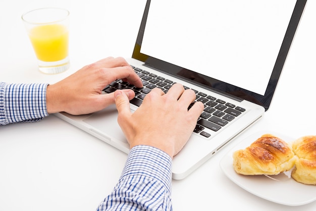 Hombre escribiendo en la computadora portátil con desayuno sobre la mesa blanca
