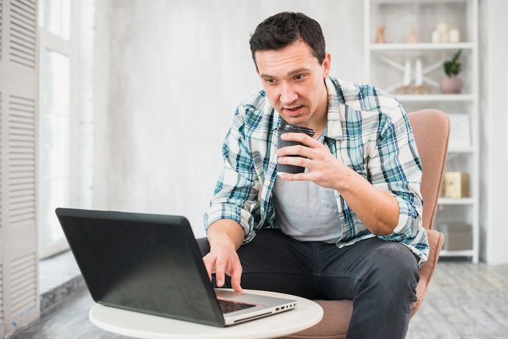 Foto gratuito hombre escribiendo en una computadora portátil y sosteniendo una taza de bebida en una silla