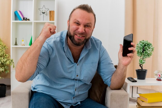 Hombre eslavo adulto alegre se sienta en un sillón manteniendo el puño hacia arriba y sosteniendo el teléfono dentro de la sala de estar