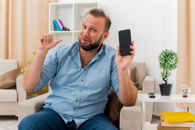 Hombre eslavo adulto alegre se sienta en un sillón sosteniendo y apuntando al teléfono dentro de la sala de estar