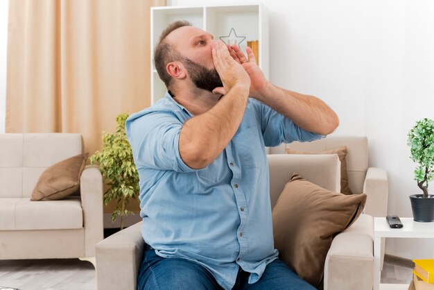 Hombre eslavo adulto alegre se sienta en un sillón tomados de la mano cerca de la boca mirando a un lado pretendiendo llamar a alguien dentro de la sala de estar