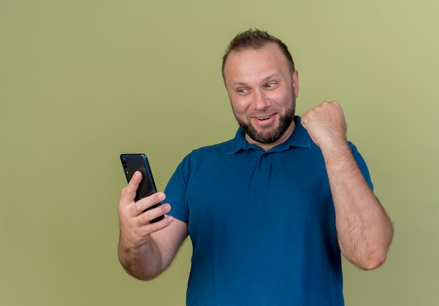 Hombre eslavo adulto alegre sosteniendo y mirando el teléfono móvil haciendo gesto de sí