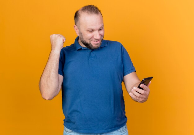 Hombre eslavo adulto alegre sosteniendo y mirando el teléfono móvil y haciendo sí gesto aislado