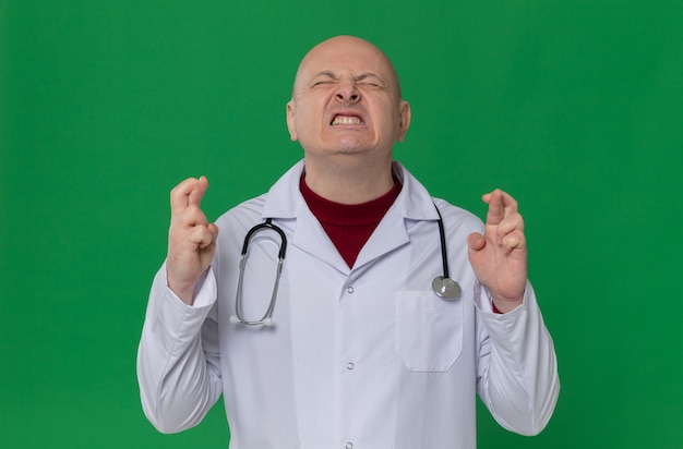 Hombre eslavo adulto ansioso en uniforme médico con estetoscopio cruzando los dedos