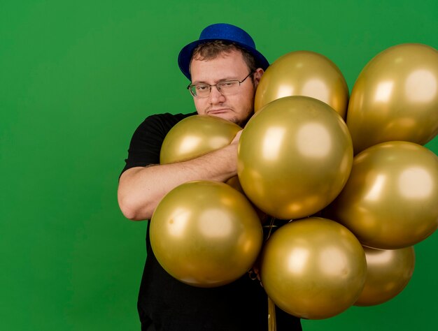 Hombre eslavo adulto codicioso en gafas ópticas con gorro de fiesta azul sostiene globos de helio