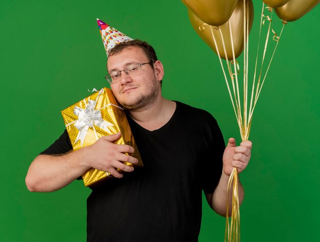 Hombre eslavo adulto complacido con gafas ópticas con gorro de cumpleaños sostiene globos de helio y caja de regalo