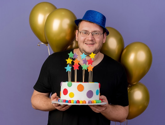Foto gratuita hombre eslavo adulto complacido en gafas ópticas con gorro de fiesta azul se encuentra frente a globos de helio tiene pastel de cumpleaños