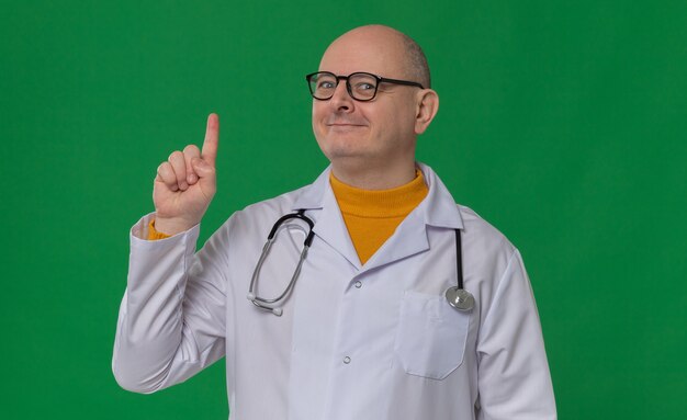 Hombre eslavo adulto complacido con gafas ópticas en uniforme médico con estetoscopio apuntando hacia arriba