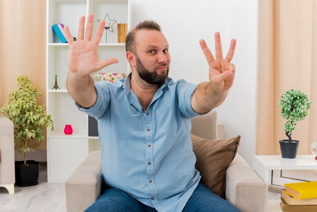 Hombre eslavo adulto complacido se sienta en un sillón gesticulando ocho con los dedos dentro de la sala de estar