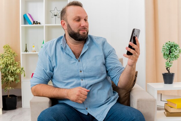 Hombre eslavo adulto complacido se sienta en un sillón sosteniendo y mirando el teléfono dentro de la sala de estar