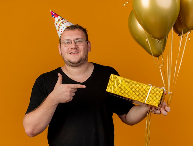 Foto gratuita hombre eslavo adulto confiado en gafas ópticas con gorro de cumpleaños sostiene y apunta a globos de helio y caja de regalo