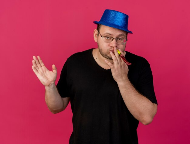 Hombre eslavo adulto confiado en gafas ópticas con sombrero de fiesta azul se encuentra con la mano levantada soplando silbato de fiesta