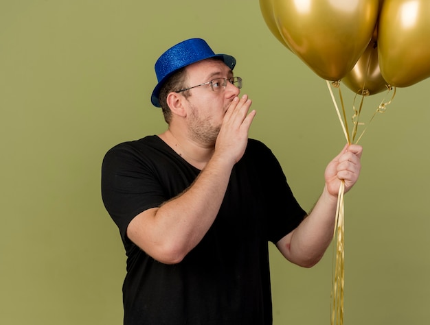 Hombre eslavo adulto confiado en gafas ópticas con sombrero de fiesta azul tiene la mano cerca de la boca mirando globos de helio